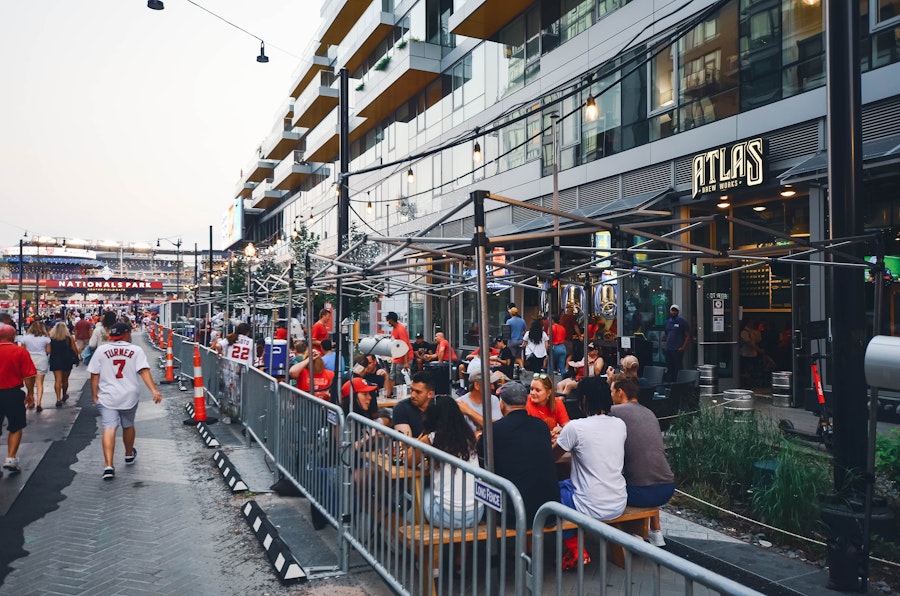 People drink beers outside of Atlas Brew Works, many of whom wear red Nationals jerseys. The stadium is visible in the background.