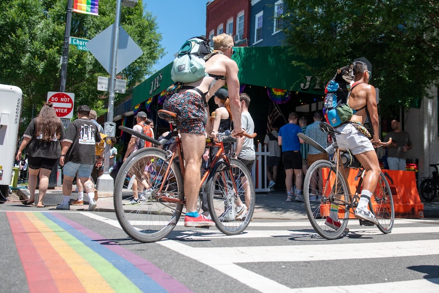 Two people ride bikes over a rainbow crosswalk during a busy festival.