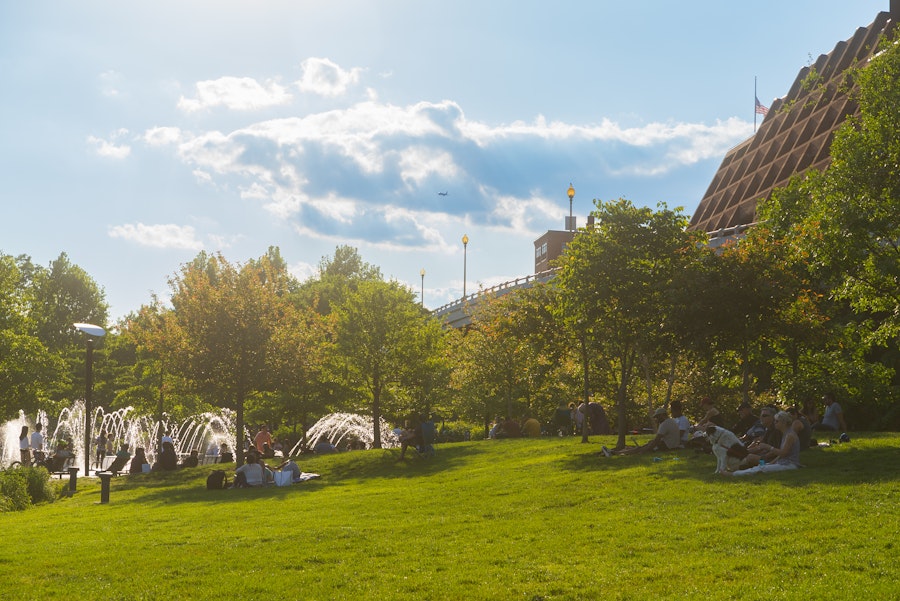 People lounge on a grassy hill near a water fountain in Georgetown on a sunny afternoon, with a modern building and bridge in the background.