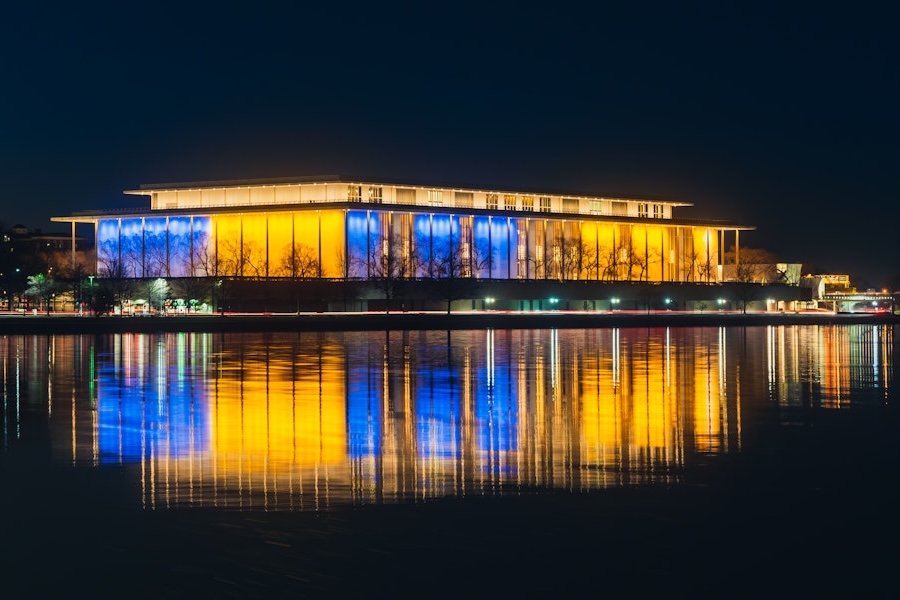 The Kennedy Center illuminated in blue and gold at night with reflections on the water.