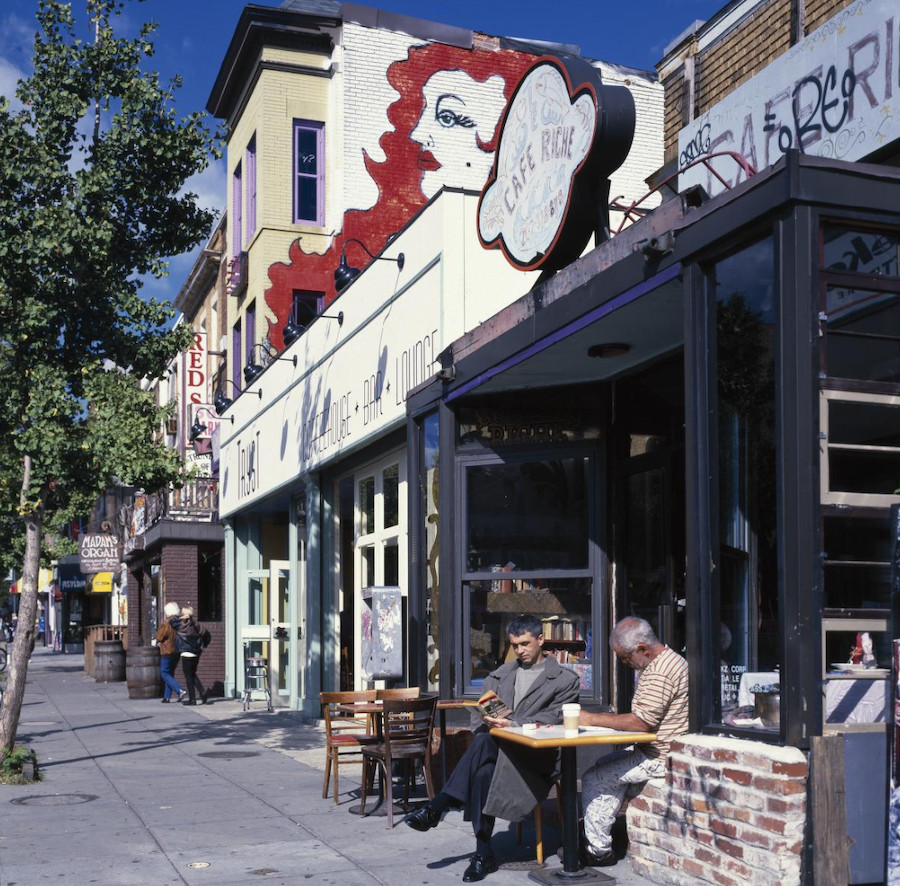 Two men sit at a sidewalk cafe table in Adams Morgan, Washington, DC with colorful murals and storefronts in the background.
