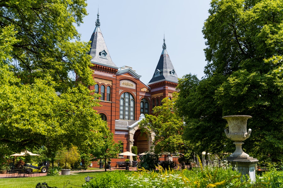 The Smithsonian Arts and Industries Building, a red-brick structure with tall spires, partially hidden by trees and garden greenery.