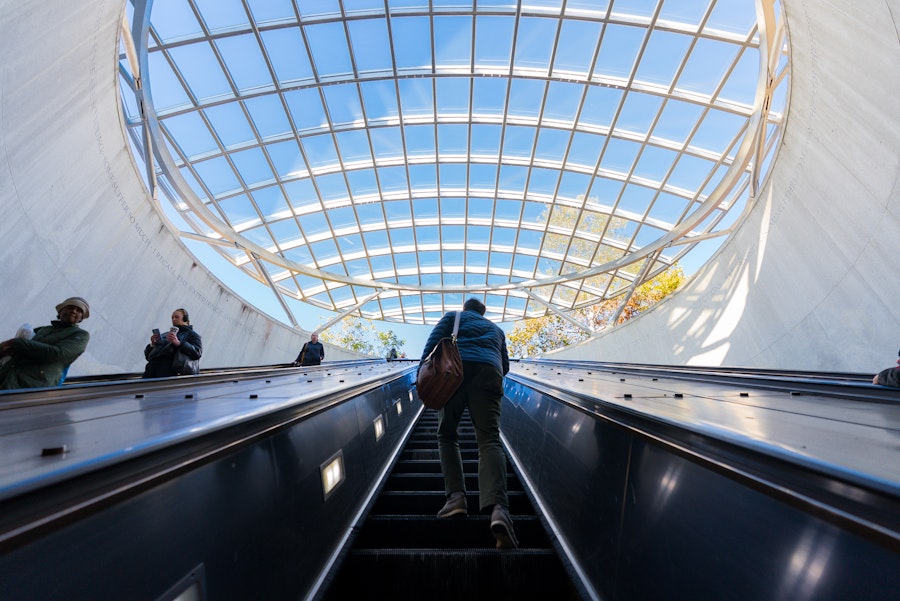 A person ascends the long escalator at Dupont Circle Metro station, with a glass dome above opening to blue sky and trees.