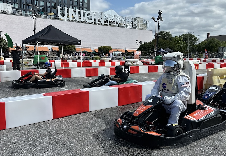 A person in an astronaut suit prepares for a go karting race, with the Union Market building and sign in the background.  