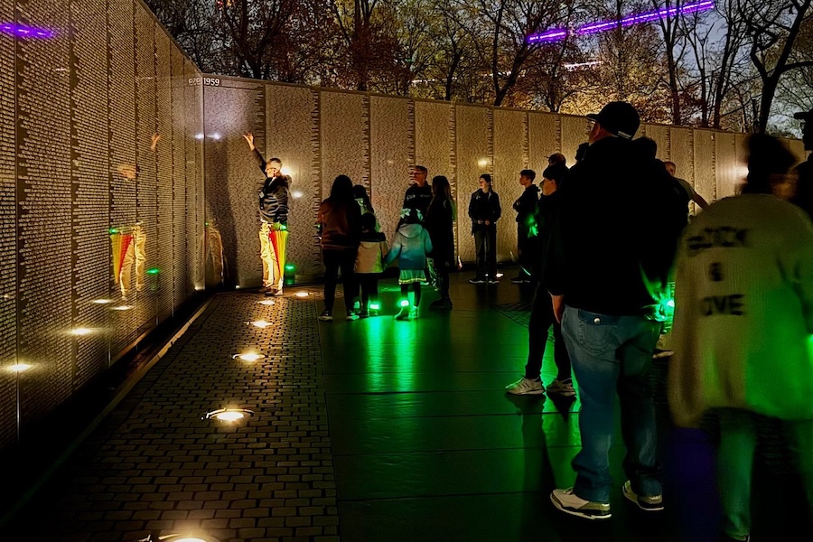 A group of travelers listen to a tour guide discuss the Vietnam Veterans Memorial in DC at night, illuminated by LED bracelets. 