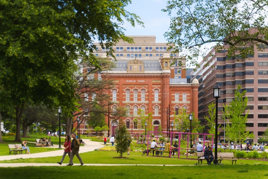 People relax and walk through Franklin Square Park with the historic red-brick Franklin School building, home to Planet Word Museum, in the background.