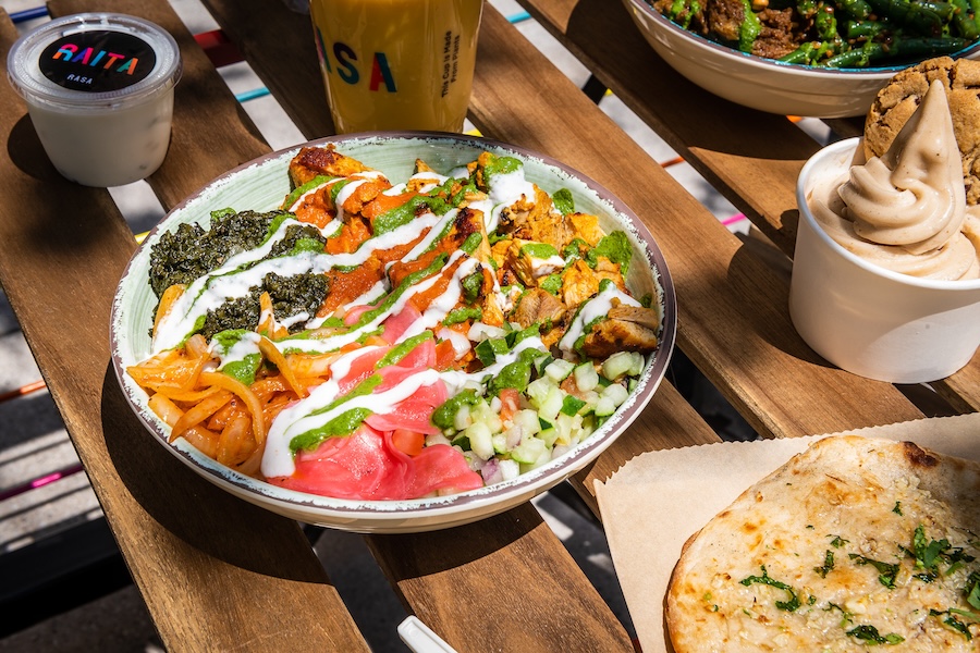 A colorful Indian-inspired bowl from Rasa with toppings like chicken, chutneys and pickled vegetables sits beside naan and soft serve.