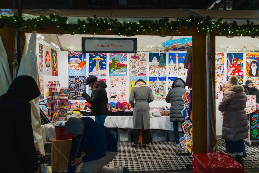 Shoppers browse colorful prints and gifts at the Naked Decor booth during a nighttime holiday market.