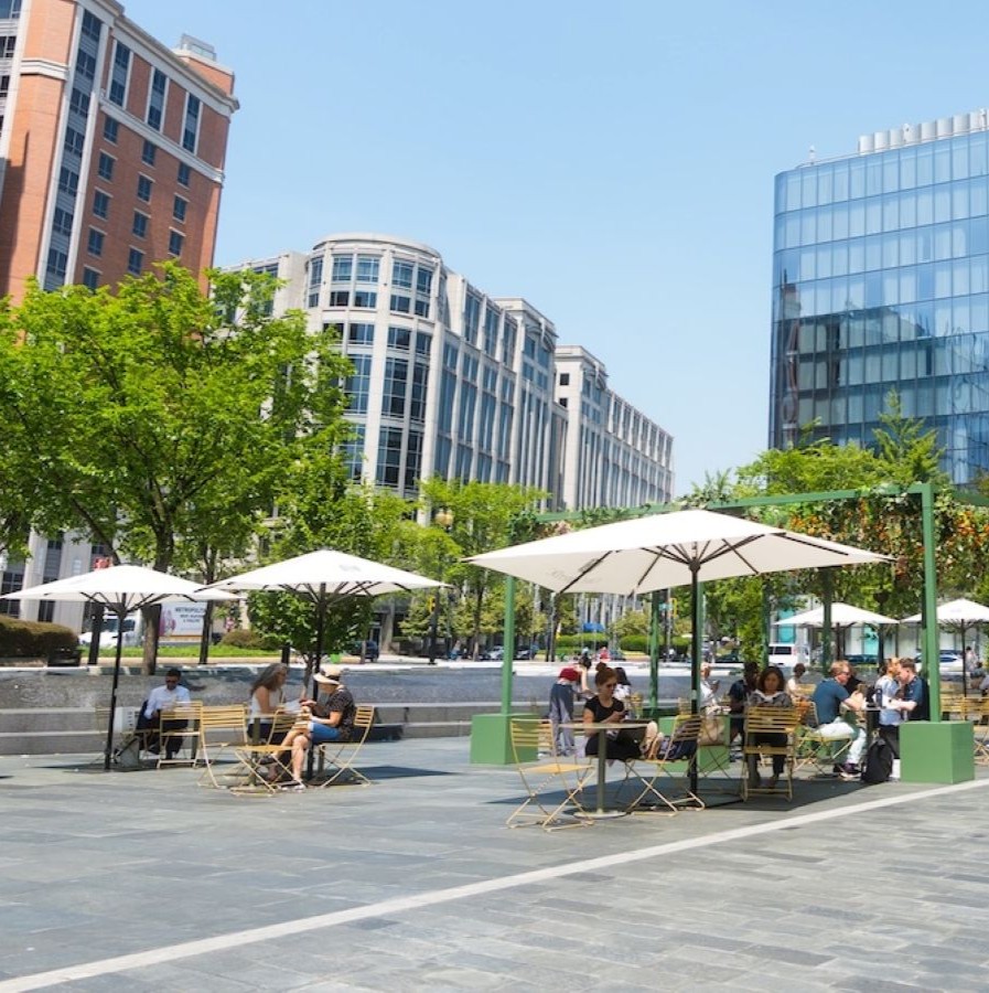 A lively outdoor plaza with people sitting under white umbrellas surrounded by modern glass buildings and leafy green trees on a sunny day.