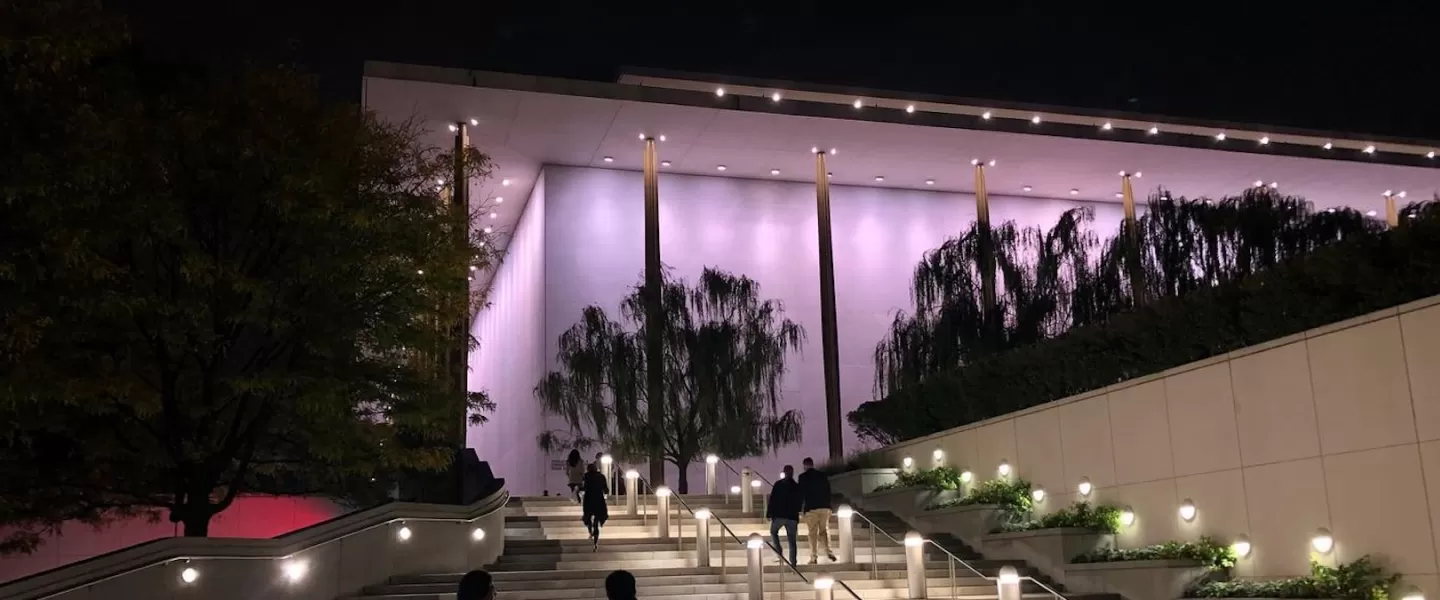 Nighttime view of the Kennedy Center's outdoor stairs, with people walking up and the building illuminated in soft purple lights.