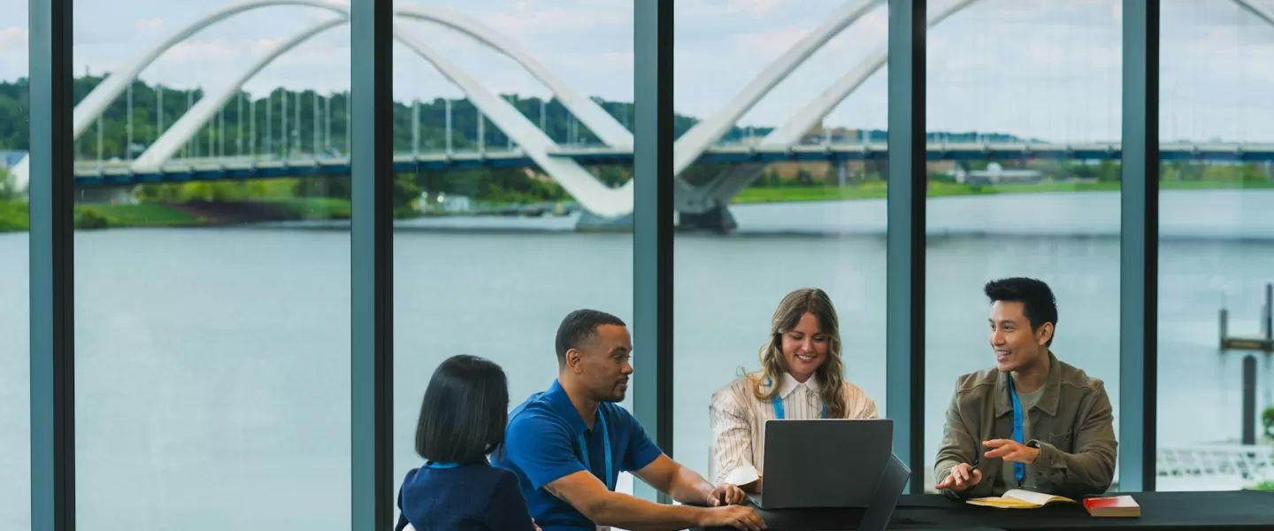 A group of colleagues collaborating around a table with laptops, set against large windows showcasing a scenic riverside view and a modern bridge.