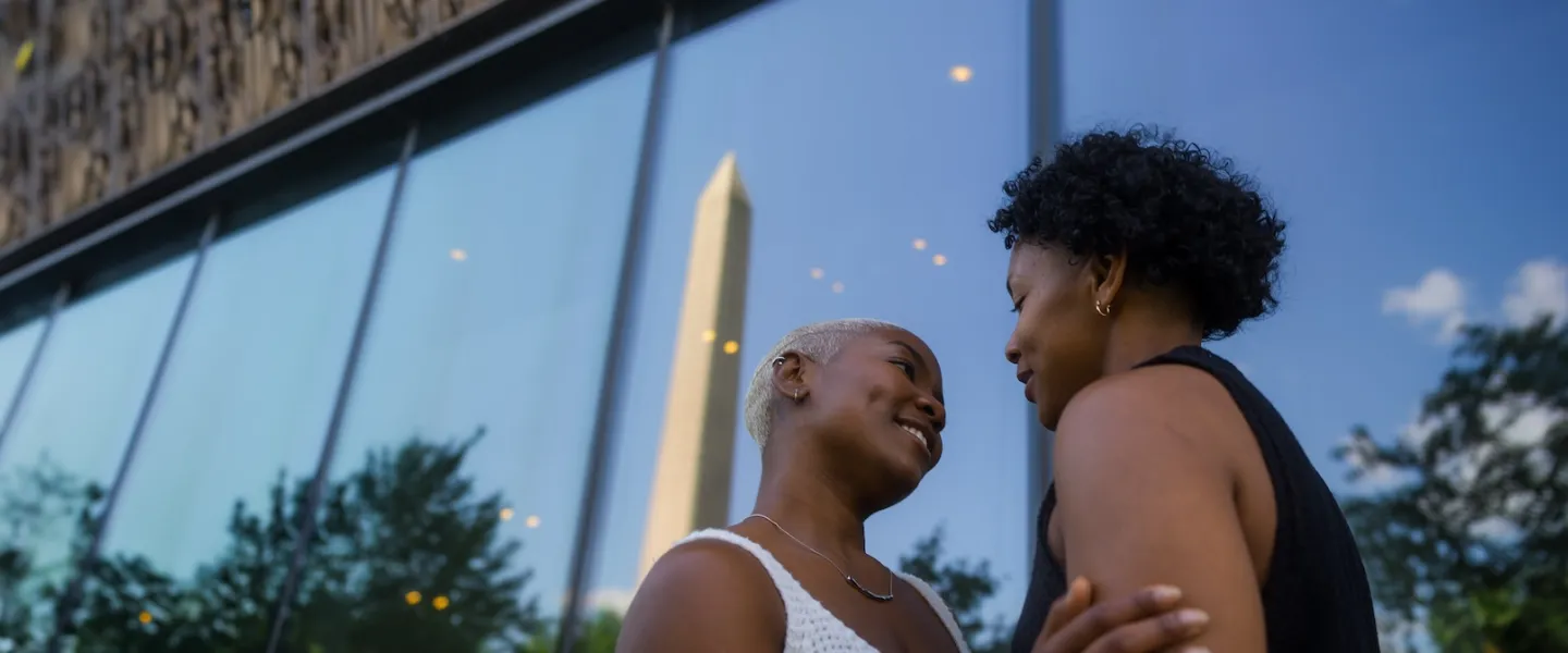 A couple shares an intimate moment in front of the Smithsonian National Museum of African American History and Culture, with the Washington Monument reflected in the glass.