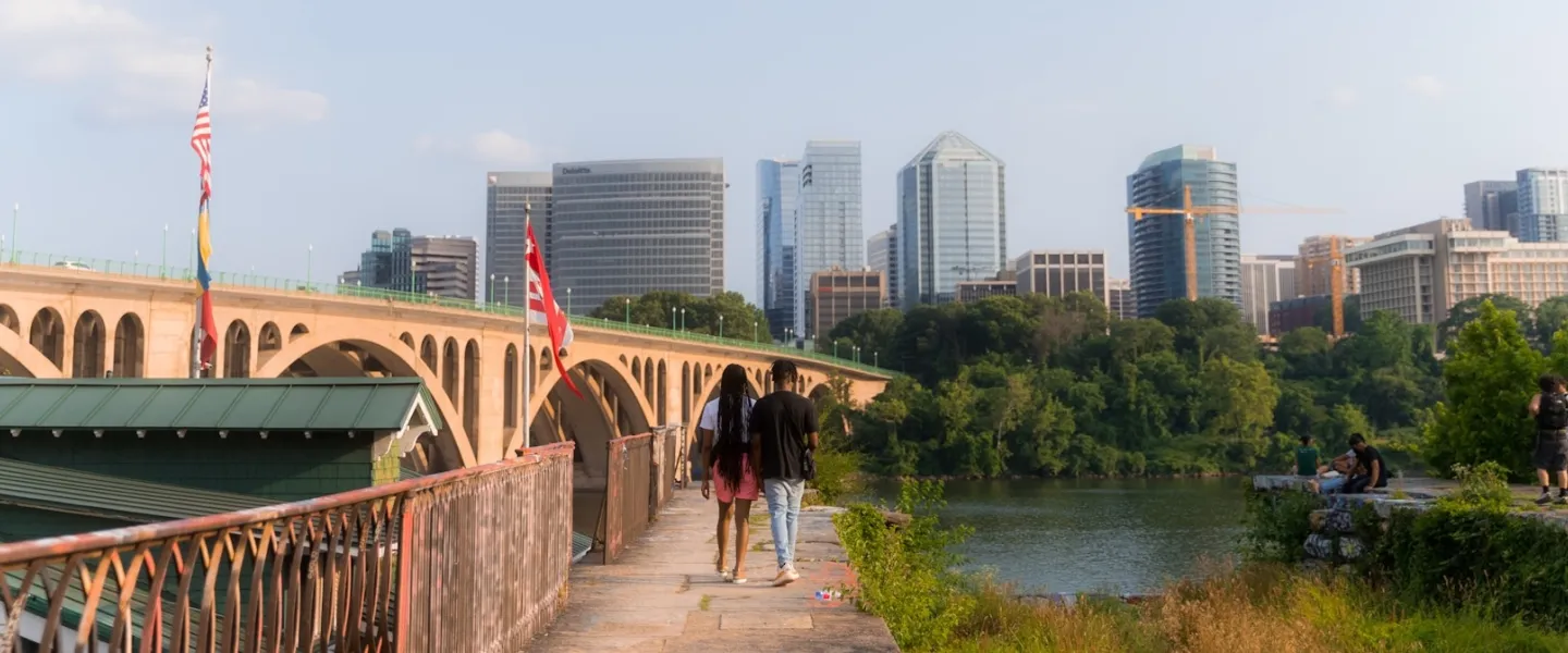 A couple walks along the Georgetown riverfront with a bridge and building of Arlington visible on the other side of the river. 