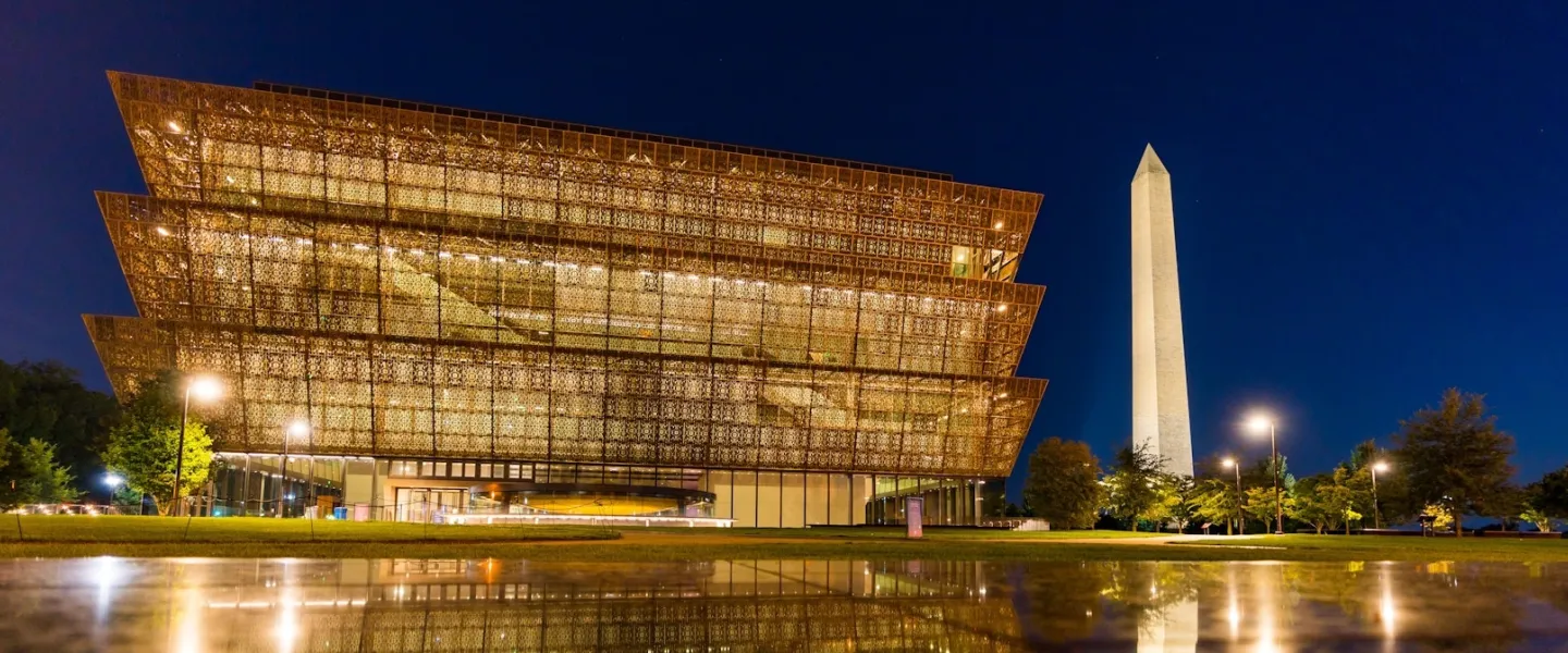 The National Museum of African American History and Culture illuminated at night with the Washington Monument in the background.