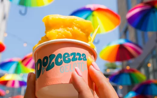 A hand holding a cup of orange gelato with vibrant, rainbow-colored umbrellas hanging overhead in the background.