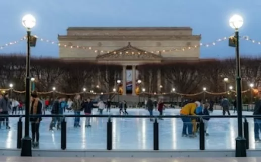 People ice skating at the National Gallery of Art Sculpture Garden rink during the evening, with string lights overhead and the museum building in the background.
