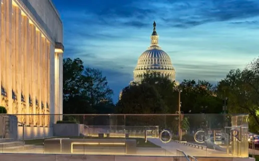 The illuminated exterior of the Folger Shakespeare Library at dusk, with the U.S. Capitol visible in the background.
