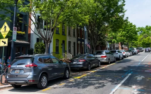 A tree-lined residential street in Georgetown with parked cars, colorful rowhouses, and pedestrians walking along the sidewalk on a sunny day.