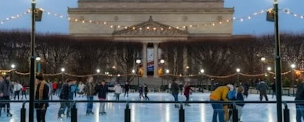 People ice skating at the National Gallery of Art Sculpture Garden rink during the evening, with string lights overhead and the museum building in the background.