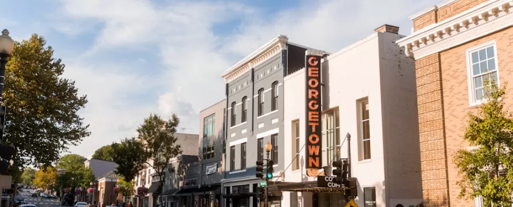 Pedestrians and cars pass by storefronts under a bright Georgetown sign on a bustling street in the historic Georgetown neighborhood of Washington, DC.