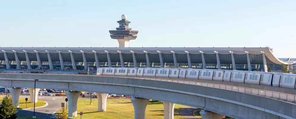 Silver Line Metro train traveling on elevated tracks with Washington Dulles International Airport terminal and air traffic control tower in the background.