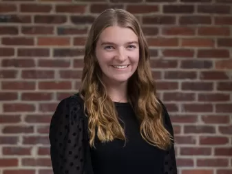 A smiling person with blonde hair wearing a black polka dot top, standing in front of a brick wall.